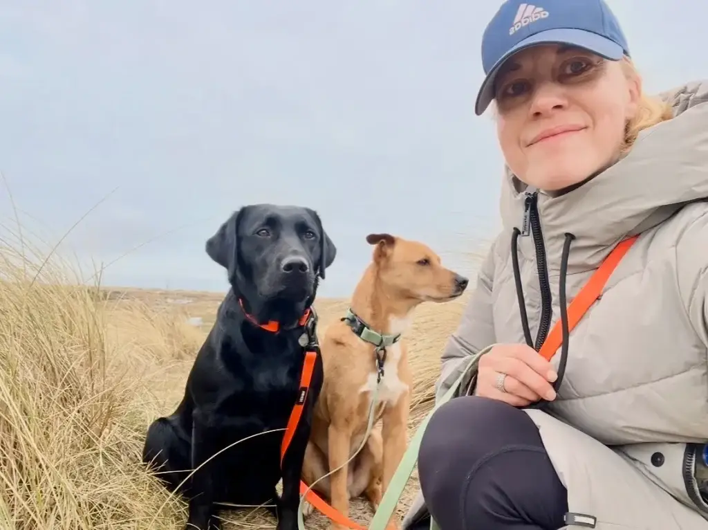 Barbara Rivers with her leashed black Labrador Pepper and her leashed Feist mix Wally sitting in the sand dunes on the Danish coast at a dog-friendly camping trip to Vejers Strand, Denmark