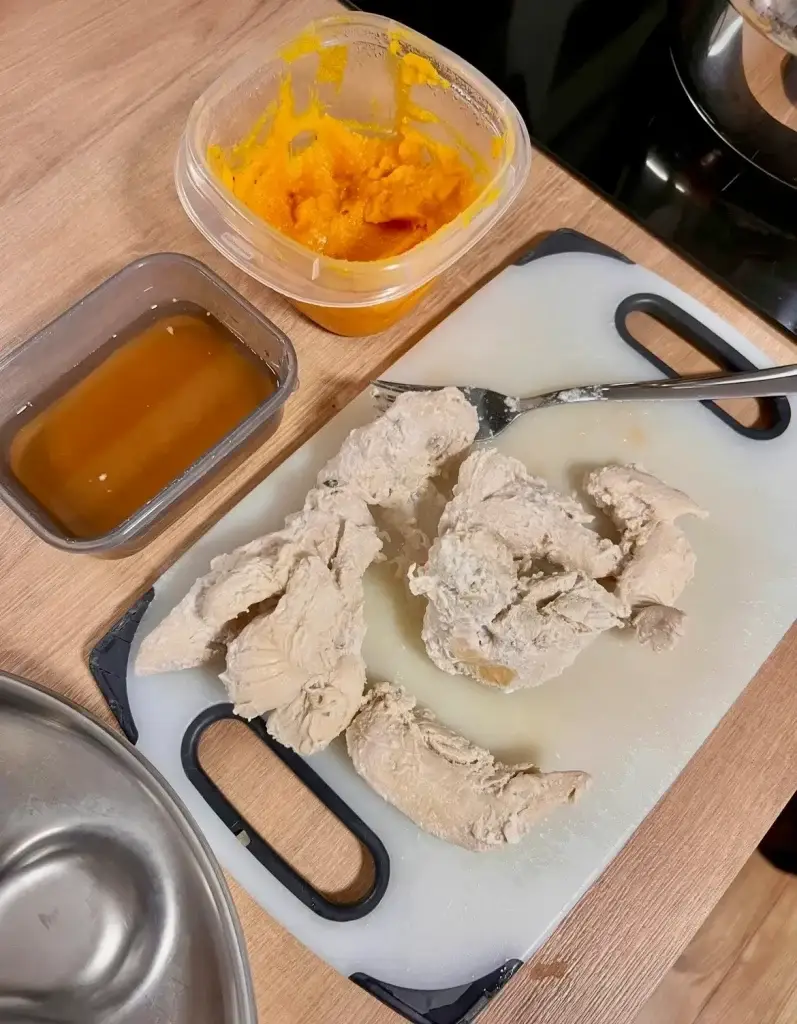 Bland diet meal for a Labrador puppy with diarrhea, showing boiled chicken breast on a cutting board next to a container of homemade pumpkin puree and a small container of slippery elm syrup, with a stainless steel dog bowl on the side